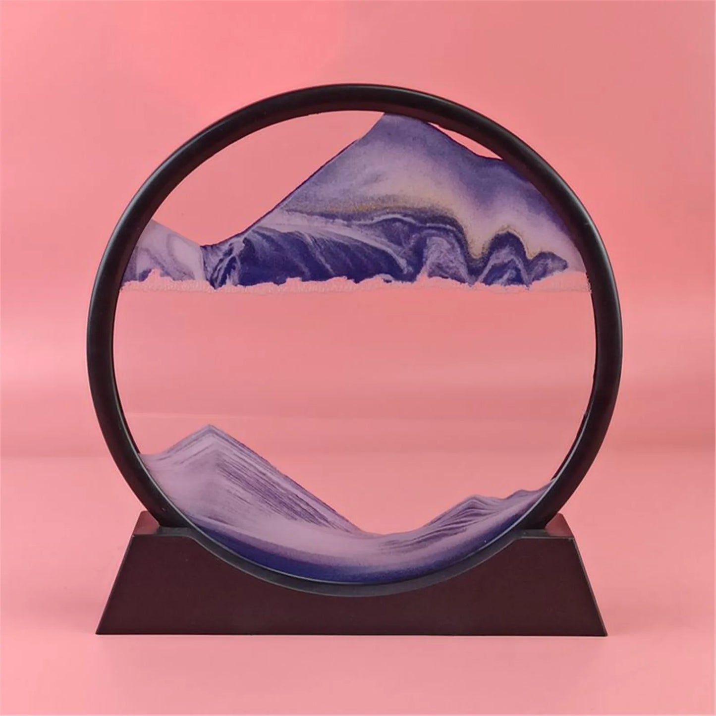 A 3D hourglass ornament with a mountain range design, containing quicksand within a circular frame placed on a flat surface.