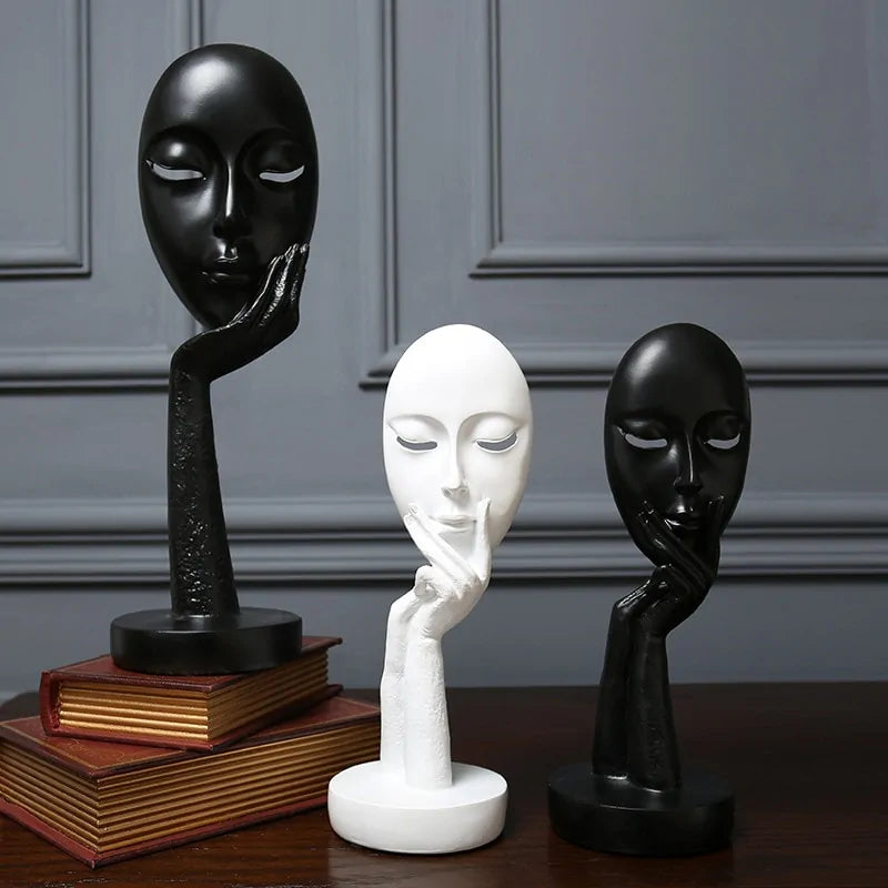 A set of three abstract sculptures in the form of faces, with one in black, one in white, and one in gray, displayed on a table with books and a shadowed background.