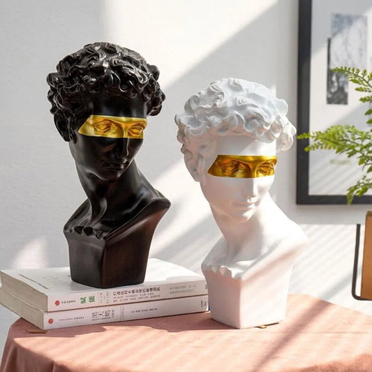 Two resin mini bust sculptures on a table, one with a black finish and the other with a white finish, both featuring abstract gold-colored eyes.